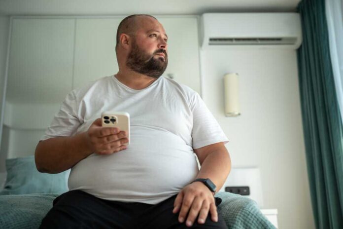 Obese,Unshaved,Man,With,Phone,And,Fitness,Tracker,Looking,At