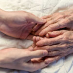 A younger persons hand gently holding an elderly persons hand on a quilted surface