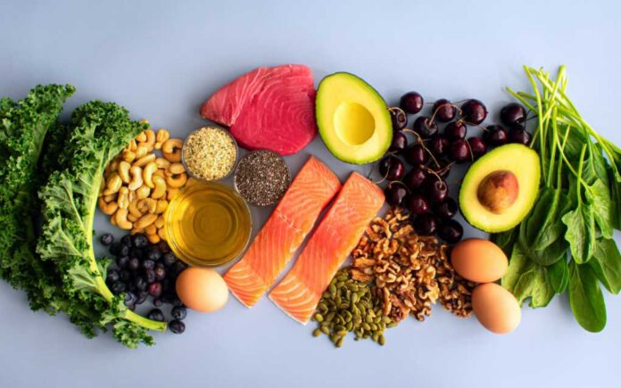 An assortment of healthy foods including fish, nuts, fruits, and vegetables arranged on a light background