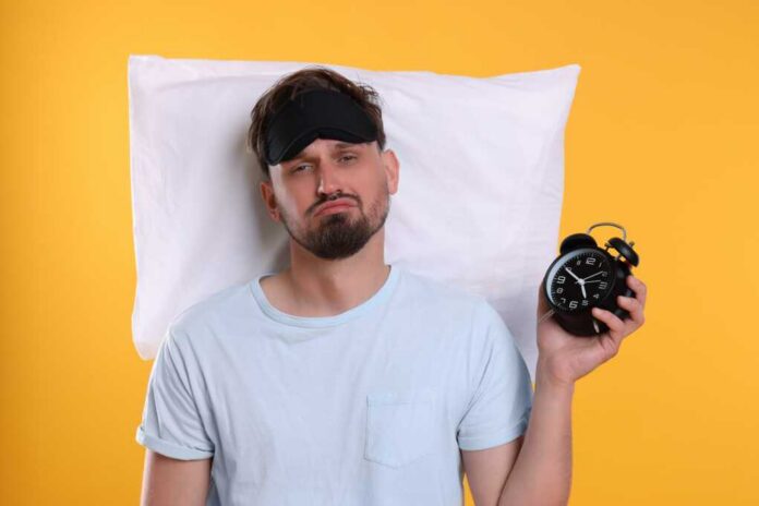 Man wearing a sleep mask holding an alarm clock with a frustrated expression