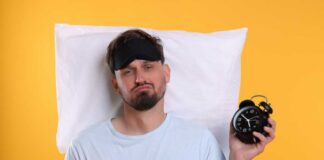Man wearing a sleep mask holding an alarm clock with a frustrated expression