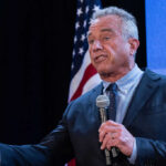 A man speaking into a microphone at a podium with an American flag in the background
