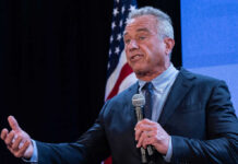 A man speaking into a microphone at a podium with an American flag in the background