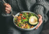 A person holding a bowl of colorful salad with avocado and greens