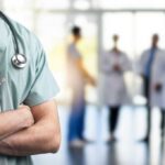 A healthcare professional in scrubs with a stethoscope, standing with arms crossed in a hospital setting