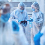Healthcare workers in protective gear discussing information on a tablet outdoors
