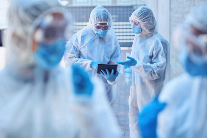 Healthcare workers in protective gear discussing information on a tablet outdoors