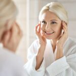 Hair Breakage or True Thinning? Smiling woman in a bathrobe applying skincare in front of a mirror