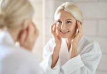 Smiling woman in a bathrobe applying skincare in front of a mirror