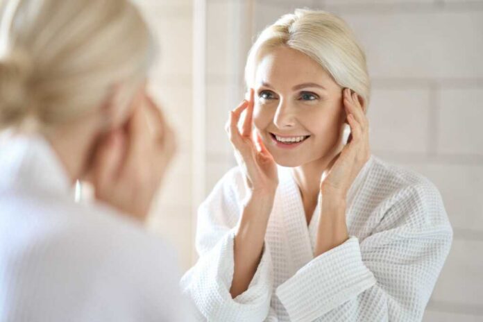 Smiling woman in a bathrobe applying skincare in front of a mirror
