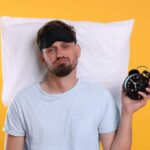 Man wearing a sleep mask holding an alarm clock with a frustrated expression