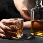 A person with their head down, holding a glass of whiskey next to a bottle