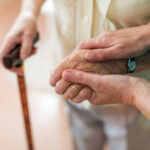 Two hands clasped together, one elderly and one younger, with a cane in the background