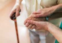 Two hands clasped together, one elderly and one younger, with a cane in the background