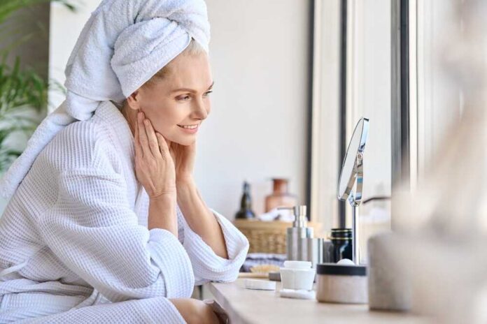 Woman in a bathrobe with a towel on her head, smiling at her reflection in a mirror