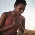 HIIT’s Surprising Link To Parkinson’s A woman in athletic wear holding her chest with a pained expression outdoors