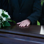 Forgotten Grievers: Sibling Loss Unseen Trauma A hand resting on a coffin adorned with white flowers during a funeral