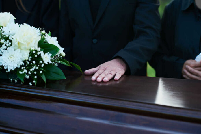 A hand resting on a coffin adorned with white flowers during a funeral