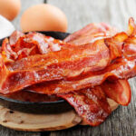 Bacon on Cancer List? — Here’s the Twist Crispy bacon strips in a cast iron skillet with eggs in the background
