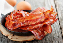 Crispy bacon strips in a cast iron skillet with eggs in the background