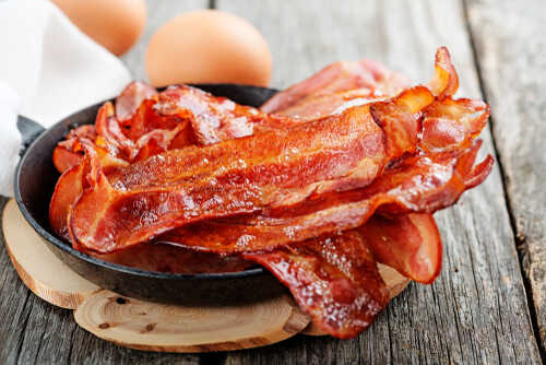 Crispy bacon strips in a cast iron skillet with eggs in the background