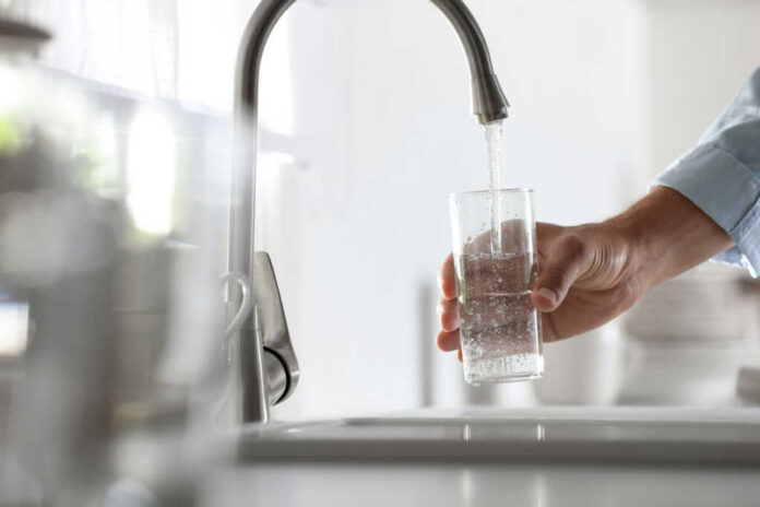 shutterstock_1928623259-1 A hand holding a glass under a kitchen faucet while water is being poured into it