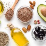 Assorted healthy fats with avocados, nuts, seeds, olives, and oils on a white background