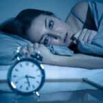 The Case Against Using Cannabis and Alcohol to Sleep Woman lying awake in bed beside an alarm clock