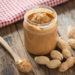 Jar of peanut butter with spoon and peanuts on rustic table