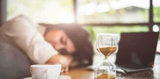 Hourglass on table with coffee cup and sleeping person in background