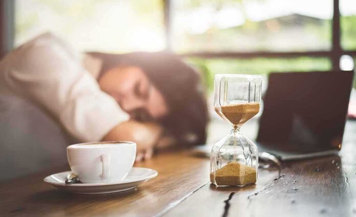 Hourglass on table with coffee cup and sleeping person in background
