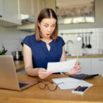 Woman in kitchen reacting to a bill at a table with laptop and calculator