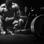 Bodybuilder kneeling beside loaded barbell in gym