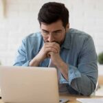 Man in glasses looking at laptop with hands clasped near mouth