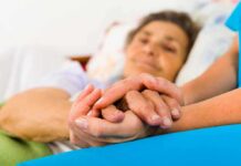 Elder Care MELTDOWN: Beds Vanish Nationwide Caregiver holding hands with an elderly patient in bed