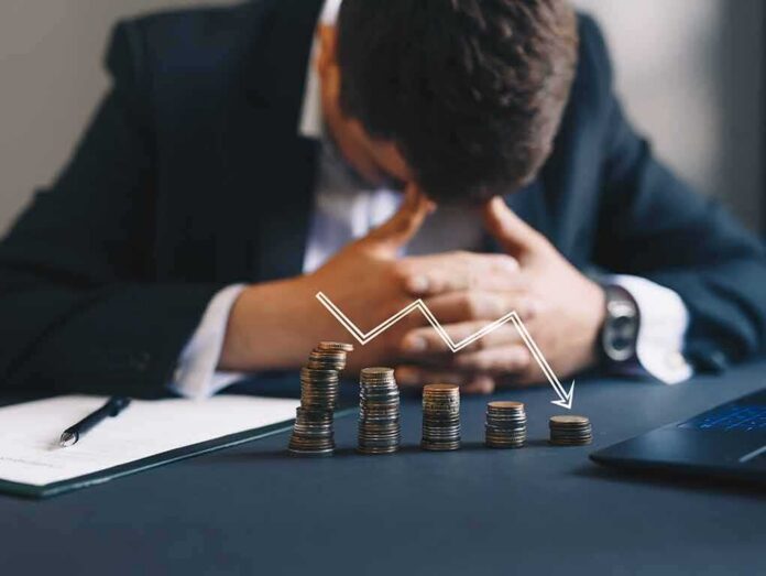 shutterstock_1694602114 Businessman with head down behind stacks of coins and downward arrow