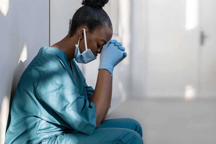 Healthcare worker in scrubs sitting against a wall with head bowed
