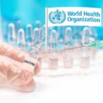 Gloved hands holding vaccine vial and syringe near WHO banner