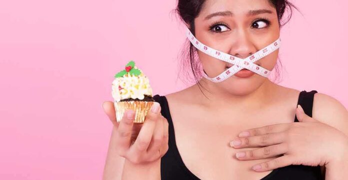 shutterstock_1870478518-1 Woman with measuring tape over mouth holding a decorated cupcake