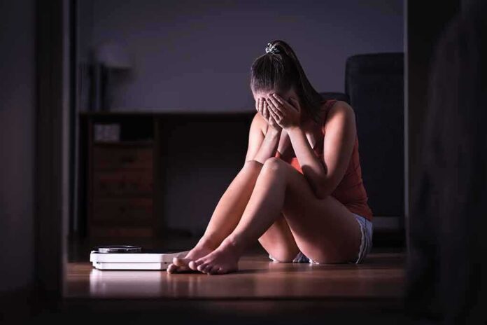 shutterstock_1291372702-1 Woman sitting on floor covering face beside a bathroom scale