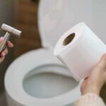 Toilet Time Trap: Doctors Sound the Alarm Hands holding a bidet sprayer and toilet paper near an open toilet